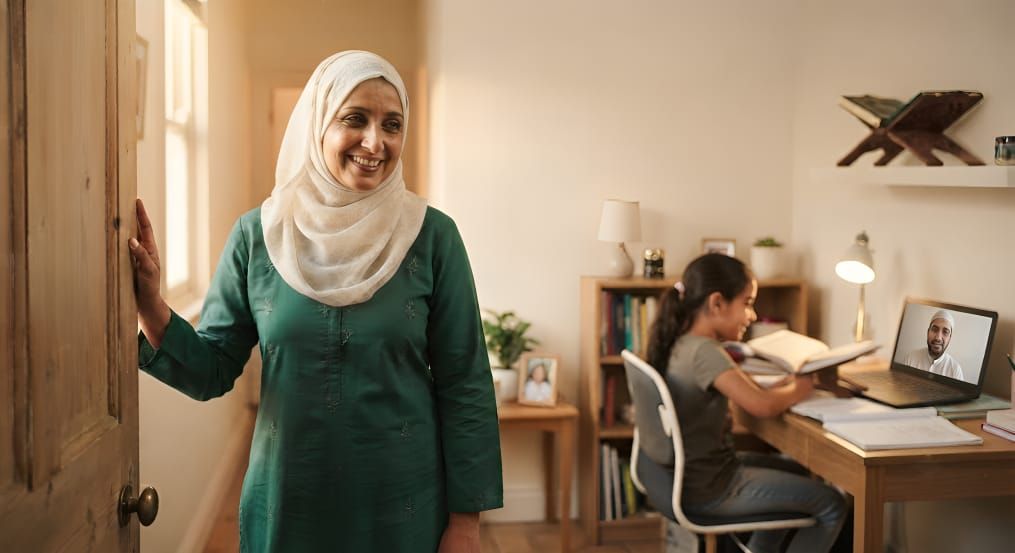 A mother stands smiling in the doorway, watching her child read confidently at a desk during a lesson with their dedicated Qurania Qari.
